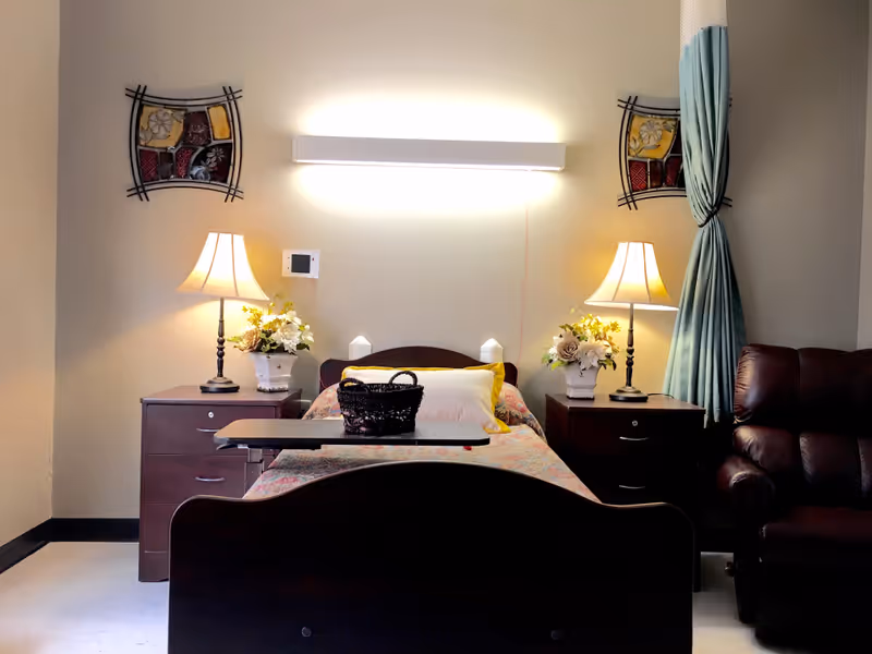 A furnished nursing facility bedroom with a single bed, overbed table, matching nightstands and lamps, floral decorations, wall art, and a recliner.