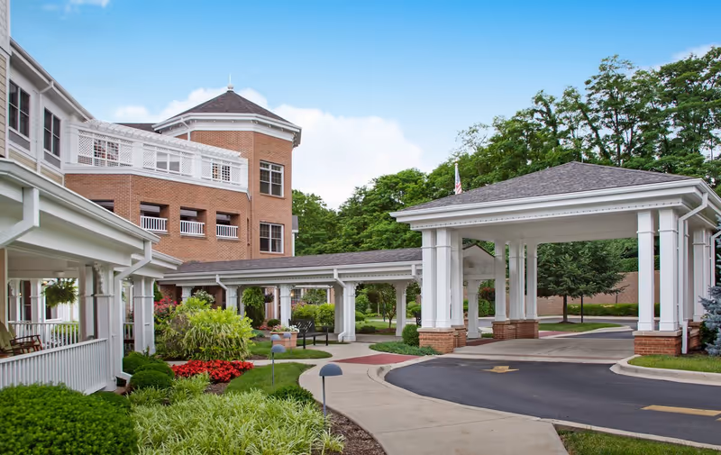 Front entrance of a brick senior living facility with a covered porte-cochere, landscaped gardens, and walkways.
