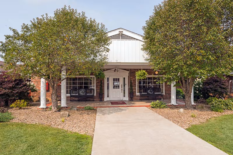 Front entrance of a single-story brick building with a covered porch, white columns, swinging benches, two trees, and a paved walkway.