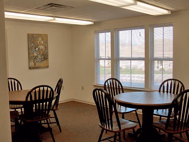 Interior room with two round wooden tables each surrounded by four wooden chairs. The room has beige walls, a carpeted floor, three large windows with blinds, and a floral painting on one wall. Fluorescent ceiling lights illuminate the space.