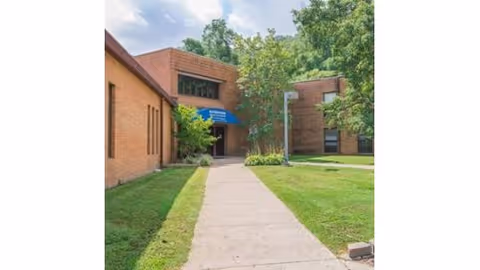 Exterior view of a brick building with a sidewalk leading to an entrance covered by a blue awning. The building is surrounded by green grass and trees under a partly cloudy sky.