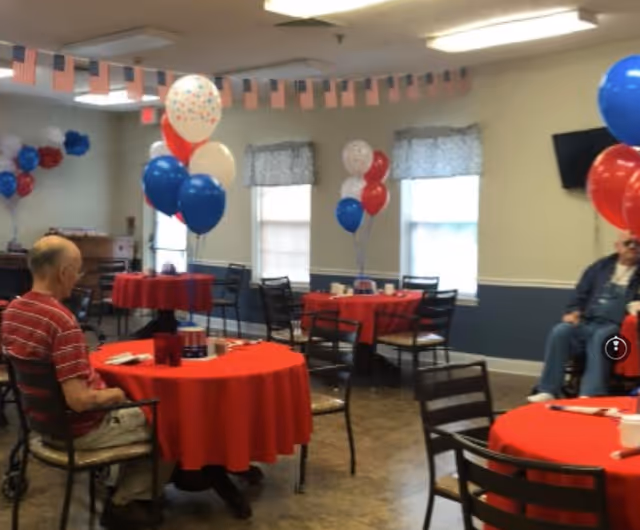 Indoor senior living dining room with red tablecloths, balloons, and residents seated at tables.