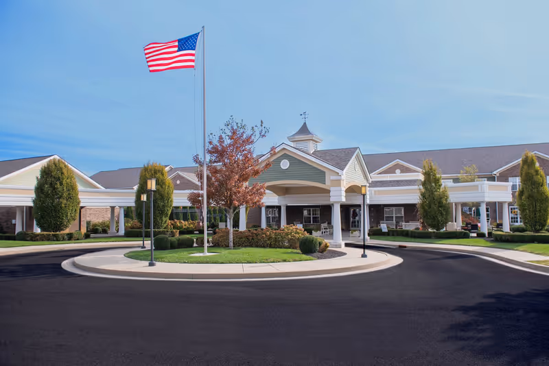 Front exterior view of Bridgepointe at Ashgrove Woods facility with a circular driveway, landscaped greenery, an American flag on a flagpole, and a covered entrance.