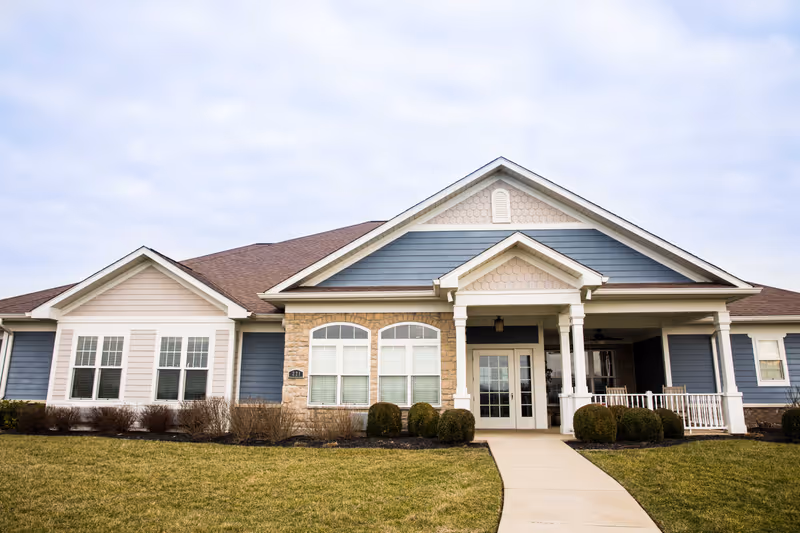 Single-story residential building with a covered porch entrance, columns, blue and beige siding, and a walkway across a front lawn.