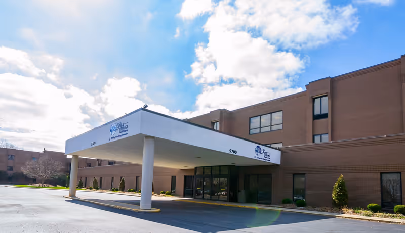 Front entrance and covered drop-off of a multi-story brick senior living building under a partly cloudy sky.