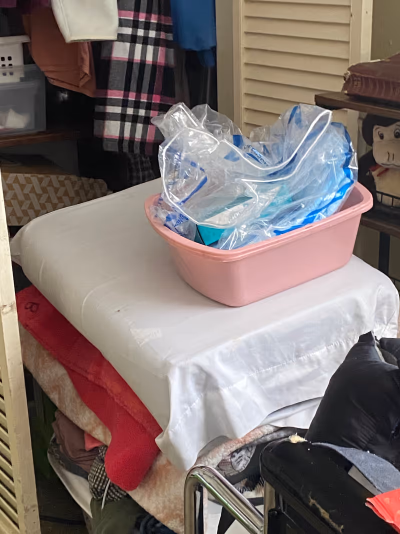 Cluttered indoor storage area with blankets, clothing, and a pink tub filled with bags.