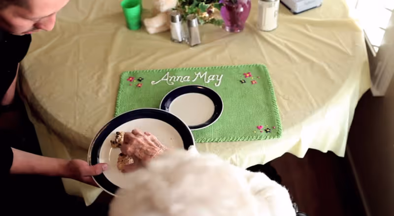 An elderly person reaching for a piece of food on a plate held by another person at a table covered with a yellow tablecloth. A green placemat with the name 'Anna May' embroidered on it is on the table along with salt and pepper shakers and a purple vase with flowers.