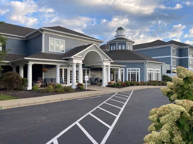 Exterior view of a senior living facility named Arcadia Louisville, featuring a blue building with white trim, a covered entrance with white columns, and a cupola on the roof. The driveway has marked parking spaces and landscaped bushes with flowers in the foreground.