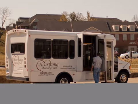 A white shuttle bus parked outside a residential area with a person standing near the open door. The bus has the logo and text 'Heartsong Memory Care' along with contact information on its side.