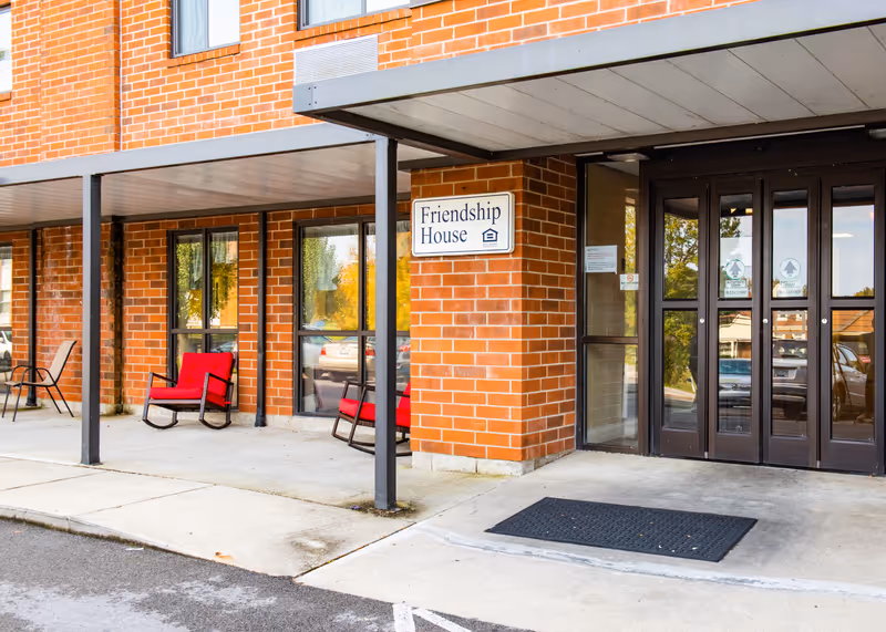 Entrance to Friendship House Corbin, a Christian Care Community, showing a brick building with a covered walkway, glass doors, and red cushioned chairs on the porch.