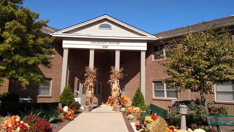 Front entrance of McCready Manor, a brick building with a white triangular pediment supported by four columns. The entrance walkway is decorated with fall-themed decorations including hay bales, pumpkins, flowers, and scarecrows. There are green benches and trees on either side of the walkway under a clear blue sky.