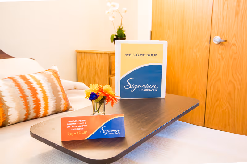 A neatly made bed with a decorative pillow and a wooden overbed table holding a small vase with colorful flowers, a welcome book, and a card from Signature HealthCARE. In the background, there is a wooden cabinet and a closed wooden door.