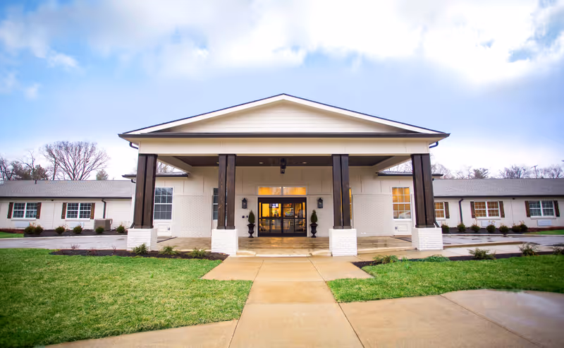 Front exterior view of Dominion Senior Living of Louisville building with a covered entrance supported by four dark wooden pillars, white brick walls, multiple windows, and a paved walkway leading to the entrance. The surrounding area has green grass and some small shrubs.