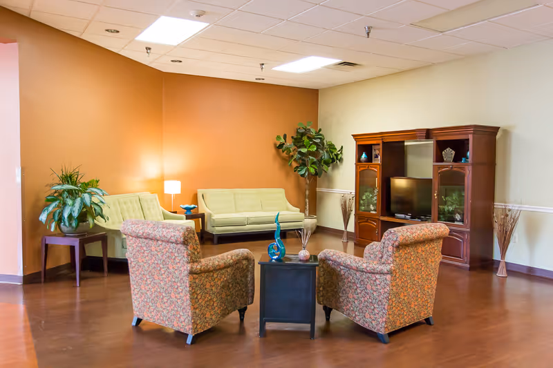 A cozy living room with two floral patterned armchairs facing a small black coffee table with decorative items. Behind the chairs, there are two light green sofas against a warm orange wall. A wooden entertainment center with a TV and decorative items is placed against a beige wall. There are also two potted plants and a floor lamp providing soft lighting.
