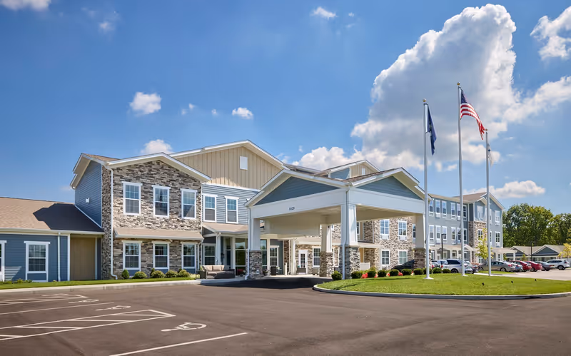 Front exterior view of Legacy Living of Florence, a multi-story senior living facility with stone and siding facade, a covered entrance, three flagpoles with flags, and a parking lot with marked spaces including handicapped spots.