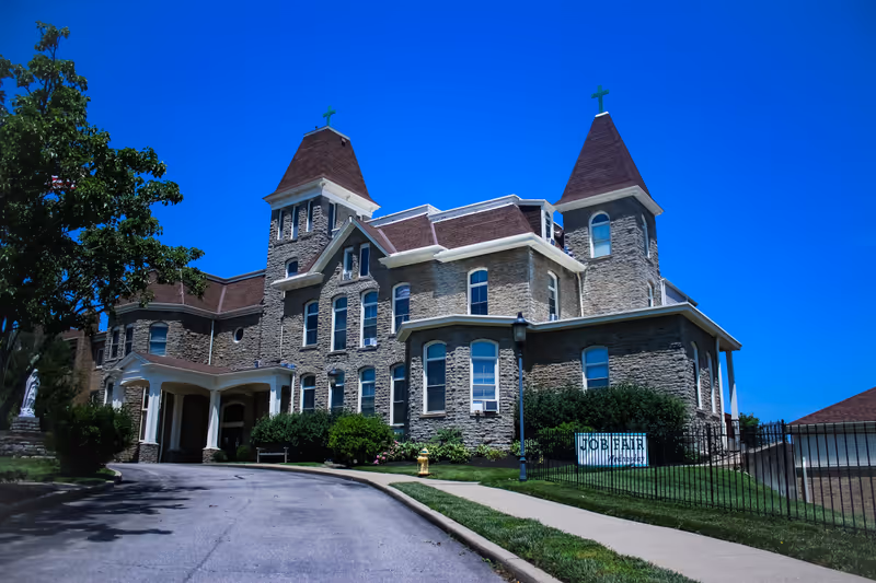 Large stone building with two towers topped with crosses under a clear blue sky, surrounded by greenery and a paved driveway leading to the entrance.