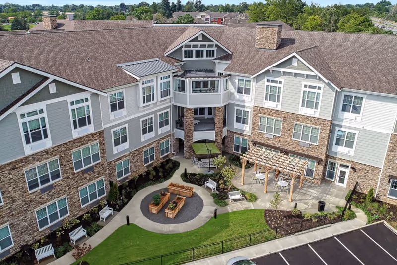 Aerial view of The Ashton on Dorsey senior living facility showing a three-story building with stone and siding exterior, multiple windows, and a landscaped courtyard with benches, raised garden beds, a pergola with tables and chairs, and a parking lot in the foreground.