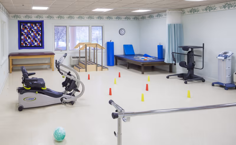 A physical therapy or rehabilitation room with exercise equipment including a recumbent bike, parallel bars, a raised platform with stairs, padded mats, and small colored cones arranged on the floor. The room has white walls, a clock, a quilt hanging on the wall, and large windows letting in natural light.