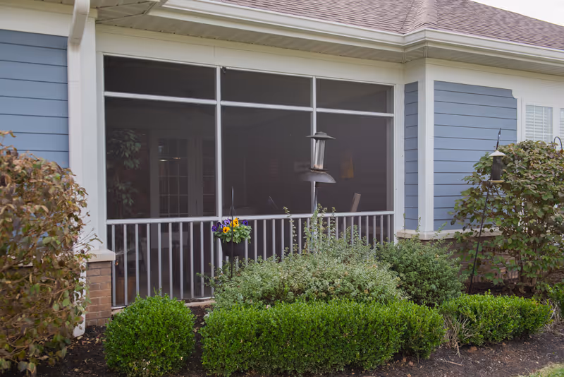Exterior view of a screened-in porch attached to a blue and white building with a sloped roof. The porch is surrounded by green shrubs and plants, with a hanging flower pot containing purple and yellow flowers. There are bird feeders visible near the porch.