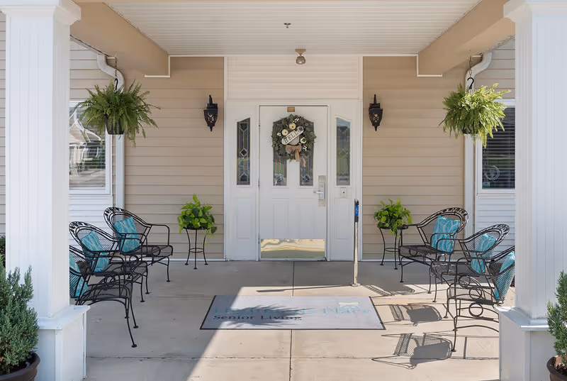 Covered entrance area of a senior living facility with a white double door decorated with a wreath that says 'Hello'. There are black metal chairs with blue cushions arranged on both sides, hanging green plants, potted plants, and beige siding on the building exterior.
