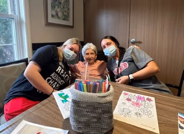 An elderly woman sitting at a table with two younger women, all wearing face masks. The table has coloring sheets and a container filled with colorful markers. They are indoors in a room with wooden walls and a window with natural light.