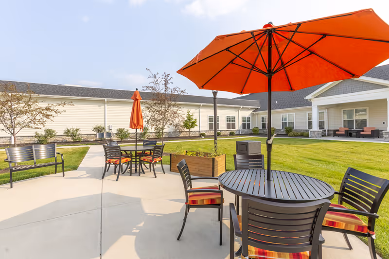 Outdoor courtyard with round patio tables, chairs with striped cushions and large orange umbrellas in front of a single-story building.