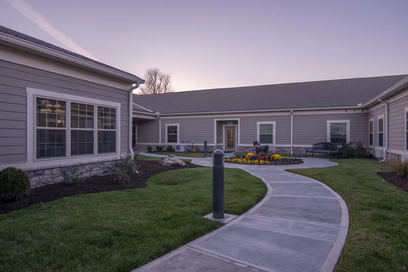 Curved concrete walkway leading through a landscaped courtyard to the entrance of a single-story senior living facility with flowerbeds and benches.