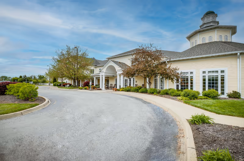 Front entrance and circular driveway of a beige senior living building with landscaped beds and trees.