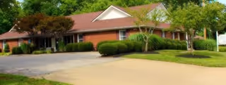 Single-story brick building with a red roof surrounded by green trees and bushes, with a paved driveway and lawn in front, under a clear sky.