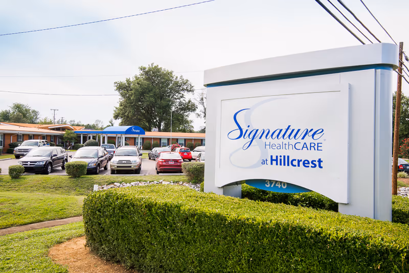 Exterior view of Signature HealthCARE at Hillcrest facility with a large sign in the foreground displaying the facility's name. Several cars are parked in front of a single-story brick building with a blue awning entrance. Green bushes and trees surround the area under a partly cloudy sky.