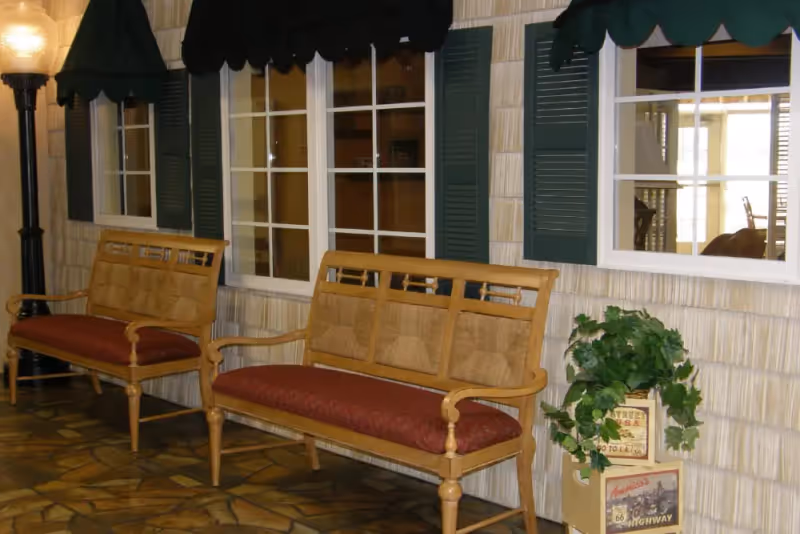 Indoor seating area with two wooden benches with red cushions beneath faux windows with green shutters and a potted plant on the right.