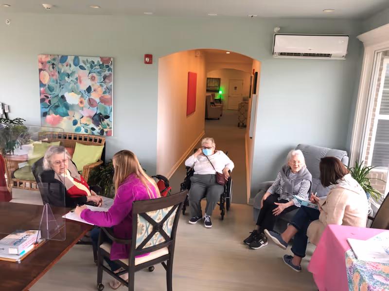 A group of elderly residents and staff talking in a bright assisted living common room, with one resident in a wheelchair.