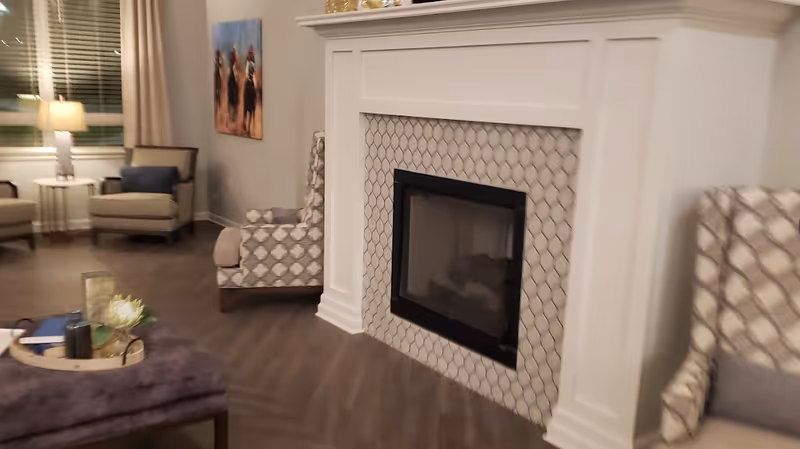 A cozy living room area featuring a white fireplace with a patterned tile surround. There are two patterned armchairs with cushions on either side of the fireplace. In the background, there are additional armchairs, a side table with a lamp, and a window with blinds and curtains. A decorative tray with a flower and other items is placed on a purple ottoman in the foreground.
