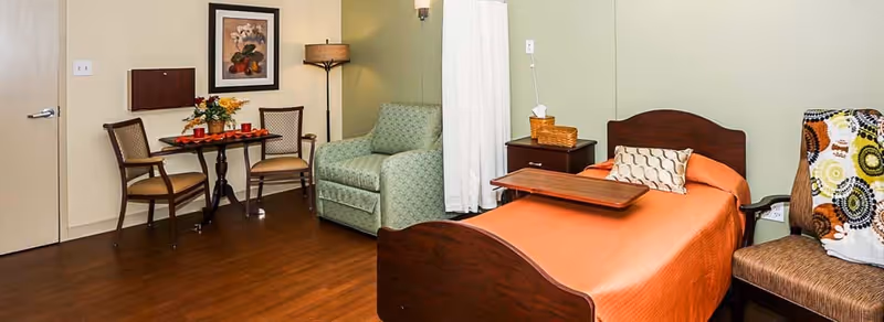 A cozy senior living room featuring a single bed with an orange bedspread and a wooden tray on top, a green patterned armchair, a small round table with two chairs, a floor lamp, and a side table with a tissue box and basket. The walls are painted light green and beige, and the floor is wooden.