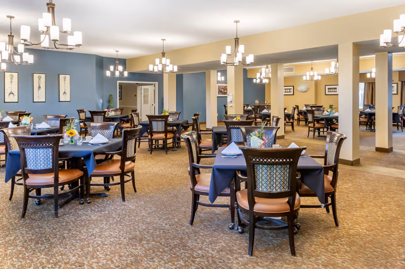 A spacious dining room with multiple round tables covered with dark tablecloths, each set with napkins, glasses, and small floral centerpieces. The room features beige walls with blue accent walls, several chandeliers hanging from the ceiling, and framed artwork on the walls. The carpet is patterned in neutral tones, and the chairs have wooden frames with cushioned seats.