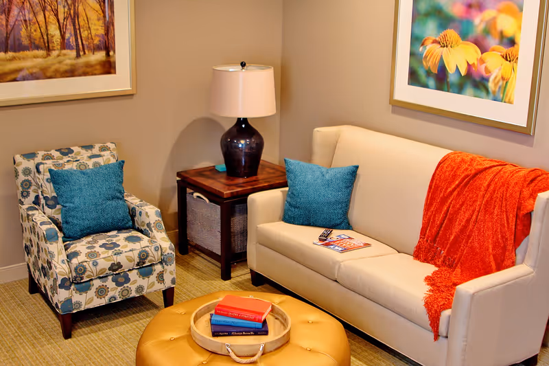 A cozy living room area with a beige sofa adorned with a blue cushion and an orange throw blanket. Next to the sofa is a floral-patterned armchair with a blue cushion. Between them is a wooden side table with a dark lamp. In front is a round, tufted ottoman with a tray holding three books. The walls feature framed artwork, one depicting a forest scene and the other yellow flowers.