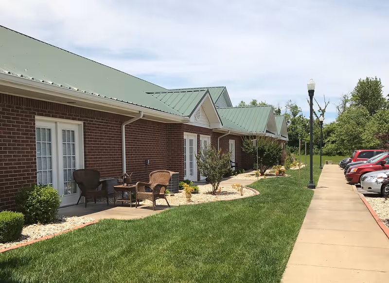 Single-story brick building with a green metal roof, front patios with wicker chairs, a grassy lawn, sidewalk and parked cars.