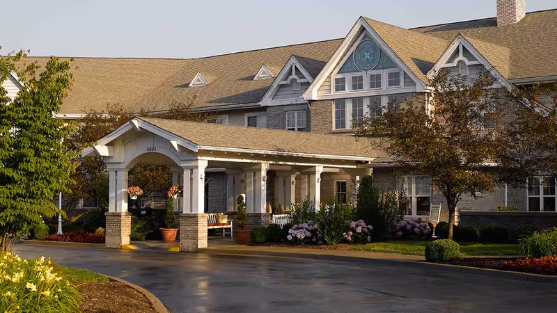 Covered porte-cochère at the front of a multi-story senior living building with landscaped flowerbeds and benches.