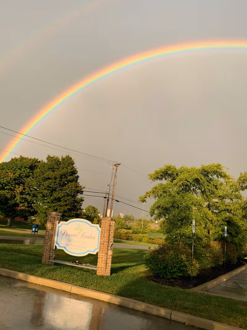 Double rainbow arches over Pleasant Meadow Assisted Living entrance with brick sign and trees.