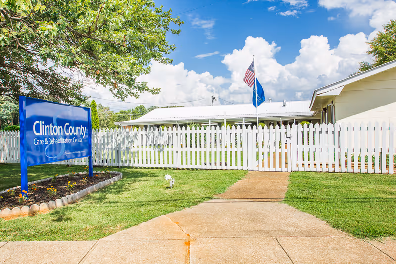 Exterior view of Clinton County Care & Rehabilitation Center showing a white picket fence, a blue sign with the facility name, a concrete walkway, green grass, trees, and two flags flying on flagpoles under a partly cloudy sky.