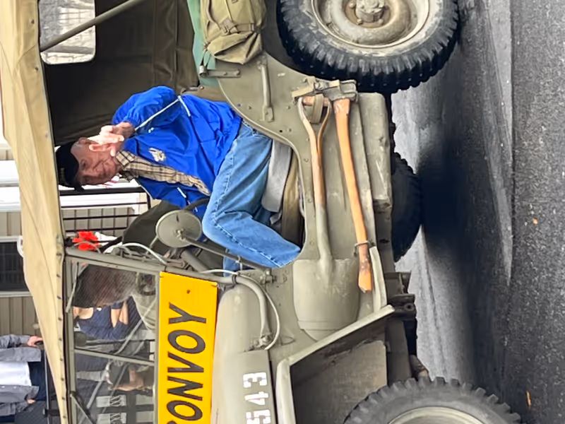Older man in blue jacket sits in a vintage jeep outside a building.