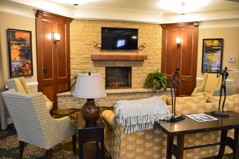 A cozy living room area with a stone fireplace and a mounted flat-screen TV above it. The room features two patterned armchairs, a yellow patterned sofa with a light blue throw blanket, a dark wooden side table with a lamp, and a console table with decorative sculptures and magazines. The walls are adorned with two colorful abstract paintings and wooden paneling around the fireplace.