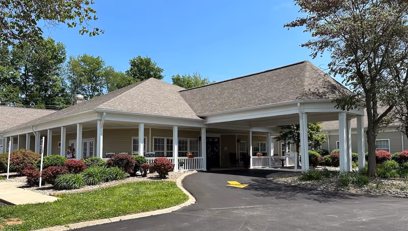 Front entrance of a single-story senior living facility with a covered porte-cochere, wraparound porch and landscaped shrub beds.