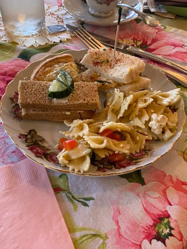 A plate with a variety of finger foods including two types of tea sandwiches, a small quiche, and a serving of bowtie pasta salad with diced tomatoes and herbs. The plate is on a floral tablecloth with a pink napkin, a glass of water, and a teacup in the background.