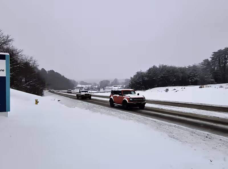 Snowy road with a red pickup driving beside trees under a gray sky