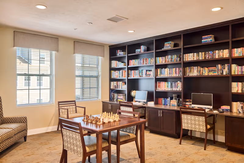 A cozy library or common area with large dark wood bookshelves filled with books, two computer workstations with monitors, a wooden table with a chessboard set up, and several patterned chairs. Two windows with blinds and beige valances let in natural light.