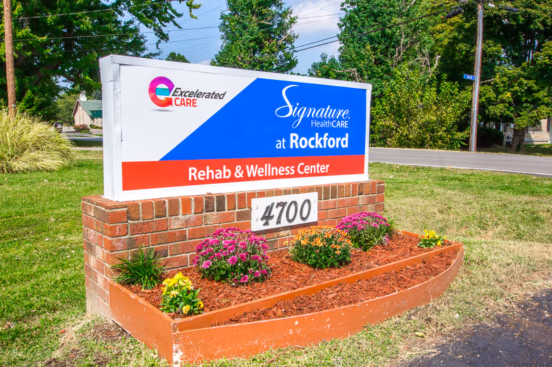 Outdoor sign for Signature HealthCARE at Rockford Rehab & Wellness Center with the address number 4700 displayed on a brick base surrounded by a flower bed with colorful flowers and mulch, set against a backdrop of green grass, trees, and a clear blue sky.