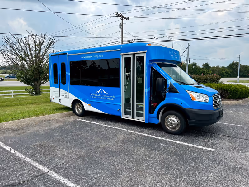A blue and white shuttle bus parked in a parking lot near a grassy area with a tree and bushes. The bus has signage for The Neighborhood at Hopkinsville, a Phoenix Senior Living Community, including their website and phone number.