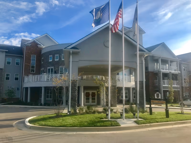 Front exterior view of Legacy Reserve at Fritz Farm building with three flagpoles displaying flags, a circular driveway, and a landscaped grassy area with small trees and shrubs.