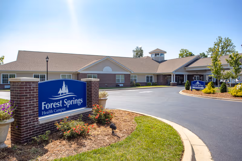 Exterior view of Forest Springs Health Campus building with a clear blue sky. The building has a beige and brick facade with multiple windows and a covered entrance. In the foreground, there is a blue sign with white text that reads 'Forest Springs Health Campus' surrounded by landscaped plants and flowers.